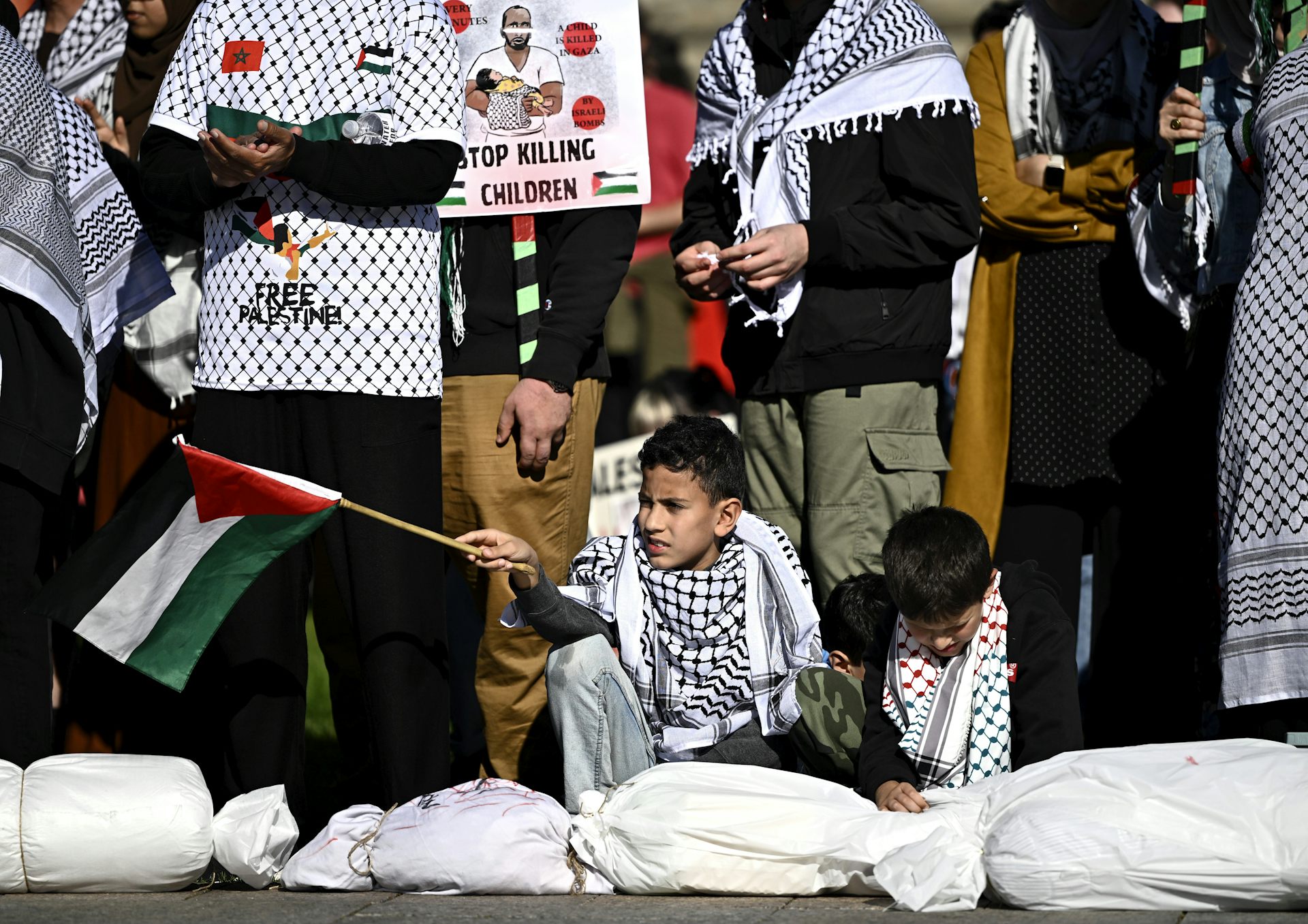 Dos niños sentados en una manifestación, uno de ellos sosteniendo una bandera palestina.