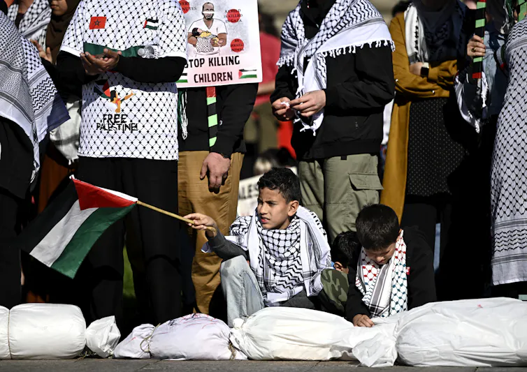 Dos niños sentados en una manifestación, uno de ellos sosteniendo una bandera palestina.