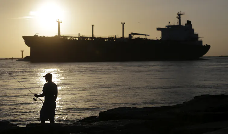 ¿Cuál es el precio de un galón de gasolina? 1 Un pescador sostiene una caña en primer plano mientras un camión cisterna pasa junto al sol.