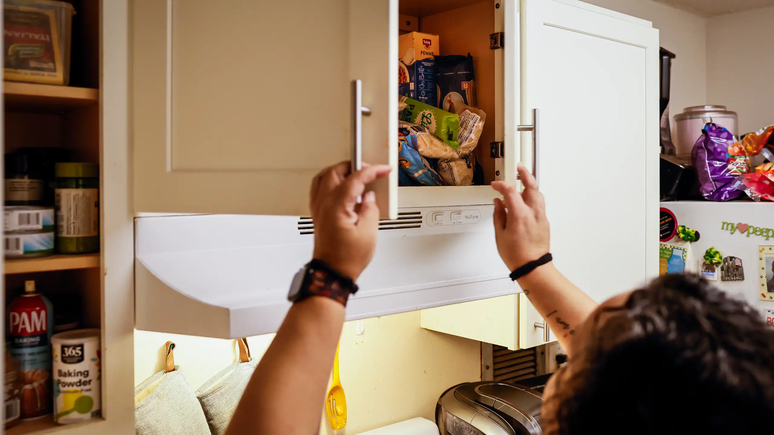 A woman holds a cabinet door open in her modest kitchen.