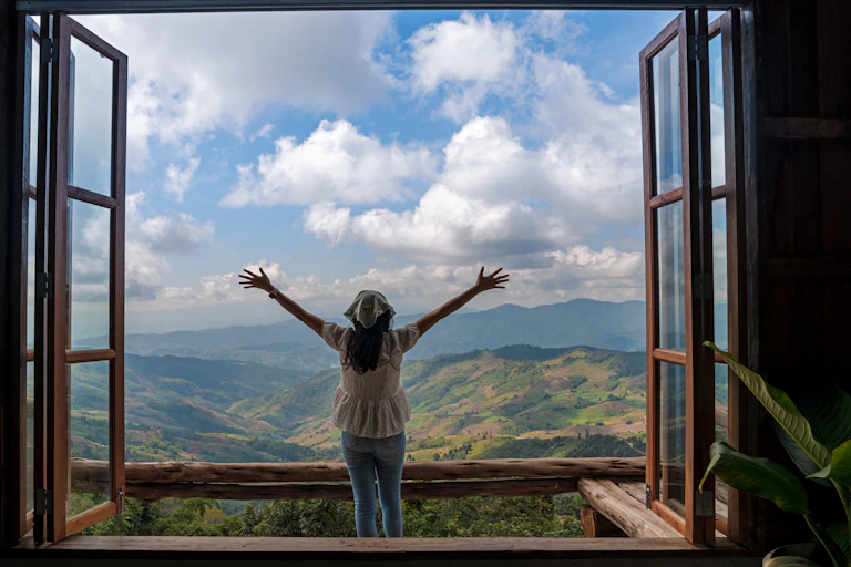 Back of a woman with arms extended up standing behind large wooden window with mountain view and cloudy blue sky