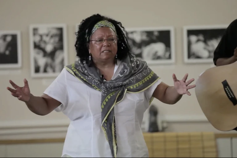 An African woman holds her arms apart in a gesture as she speaks alongside a man playing the guitar.