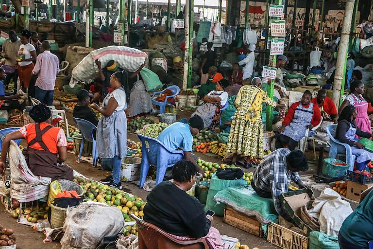Women seated behind piles of fresh produce on the ground in a marketplace