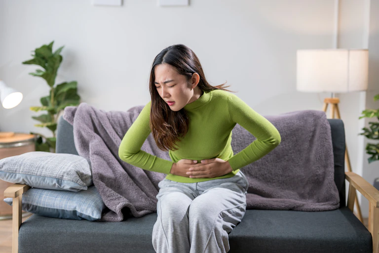A woman sits on her couch, holding her stomach in pain.