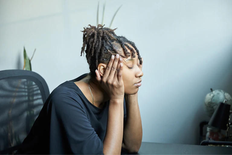 Woman sits at table with eyes closed and fingers on temples