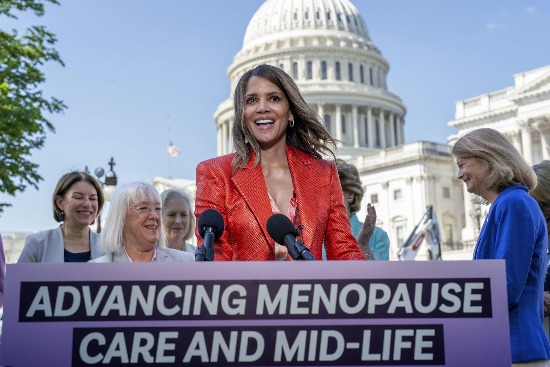Woman in red blazer stands at podium with other women and the U.S. Capitol building behind her