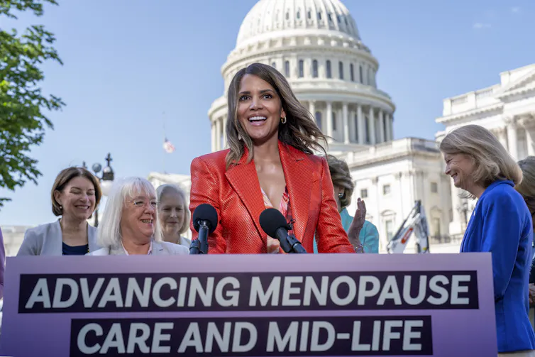 Woman in red blazer stands at podium with other women and the U.S. Capitol building behind her