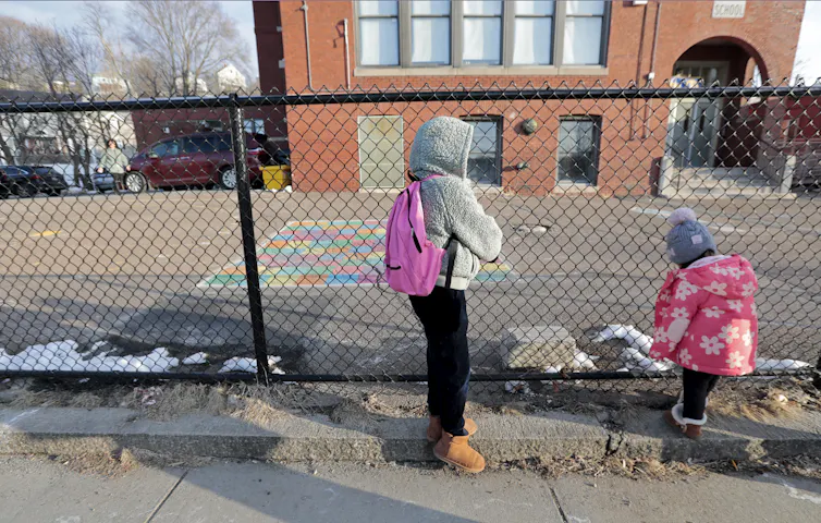 Two young girls stand near a fence outside of a brick building. They're wearing jackets and hoods and hats that cover their faces as they look away from the camera and toward the building.