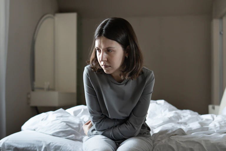 A sad woman sits on her bed looking tired or in pain.
