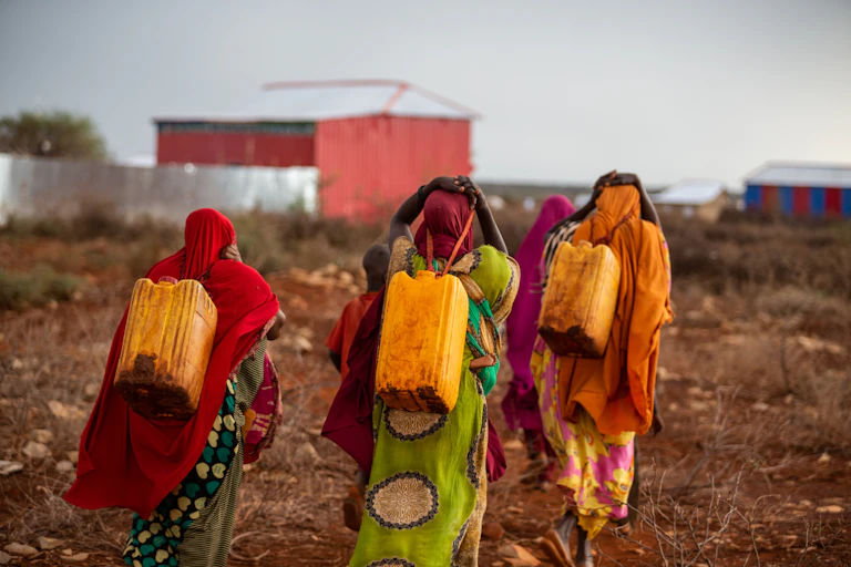 Three women in colourful clothes are carrying water canisters on their backs