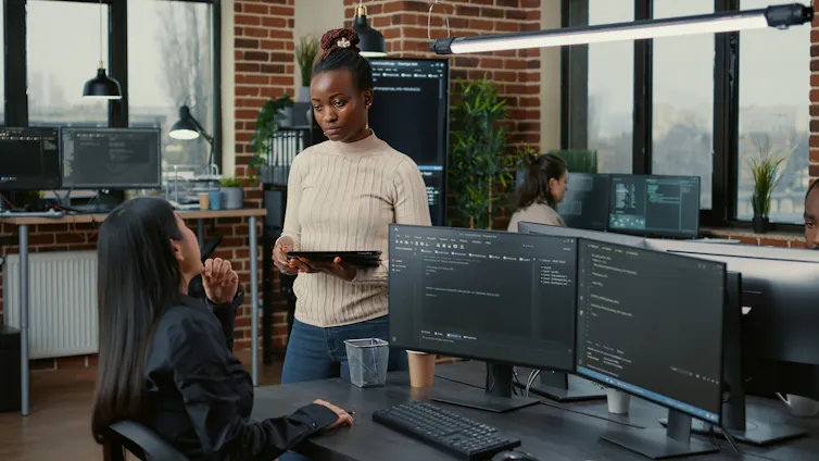 Two workers in a tech-heavy office having a conversation while one is standing and the other is sitting at her desk.