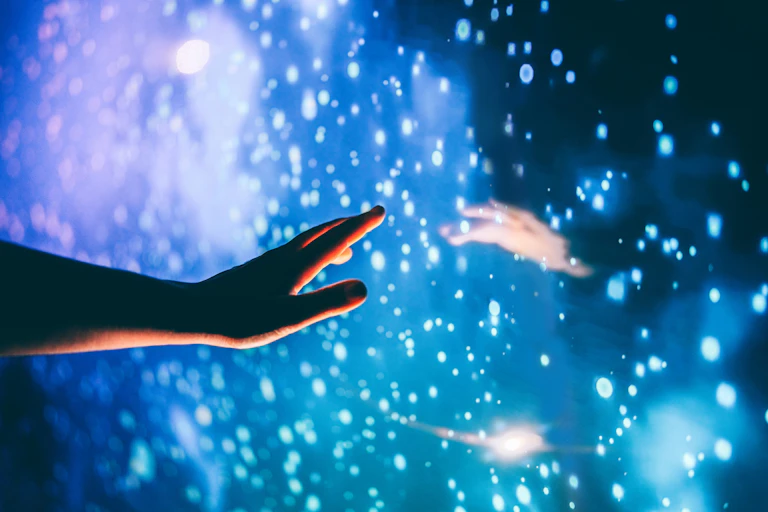 Hand reaching out to a dark, glowing surface with white circles of light, a reflection of another hand reaching back