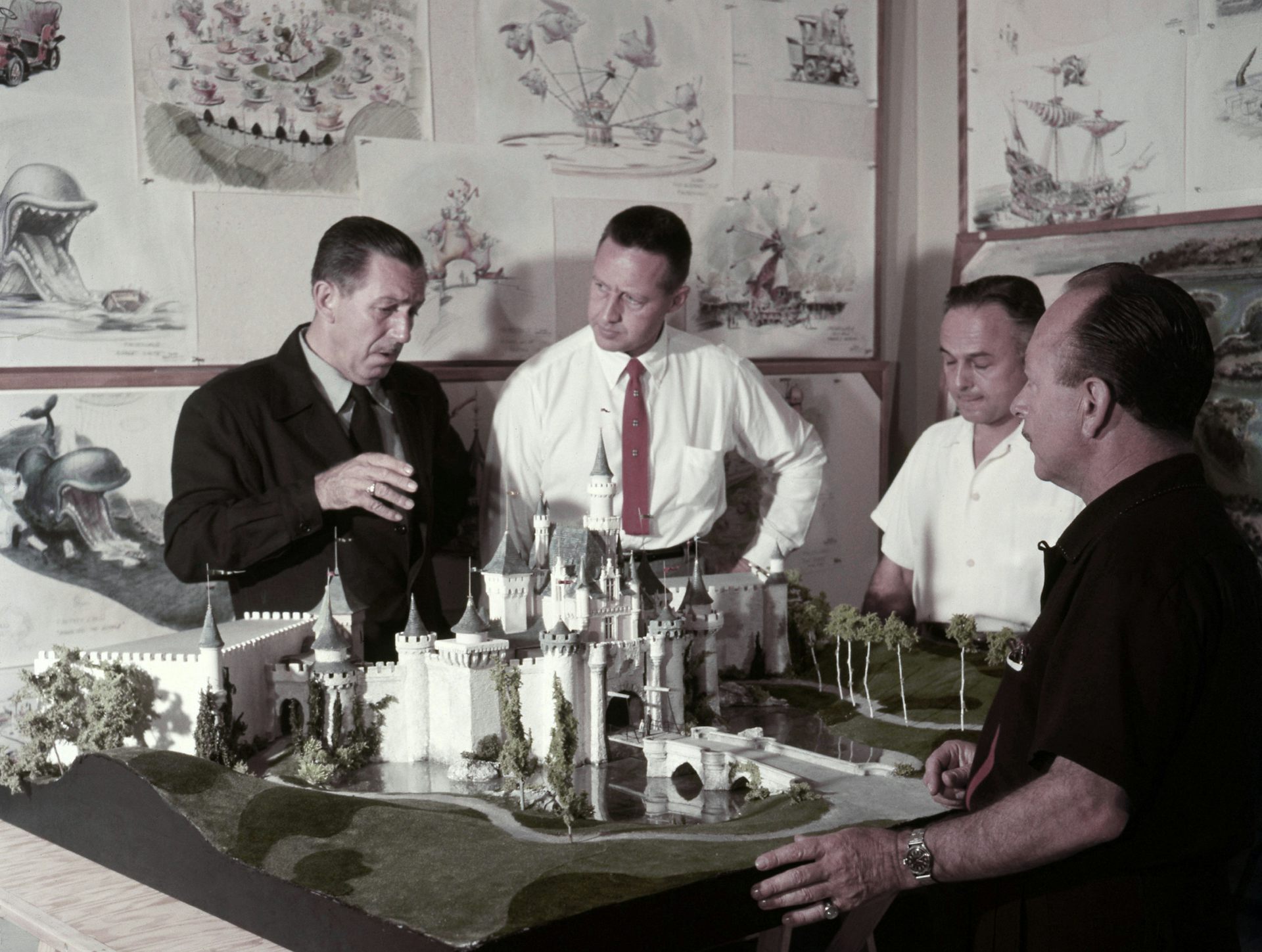 A group of men gathering around a model of a castle and chatting with one another.