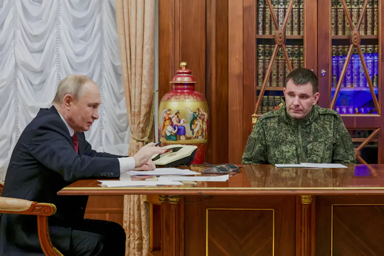 A balding older man at a wood table next to a younger man in battle fatigues.