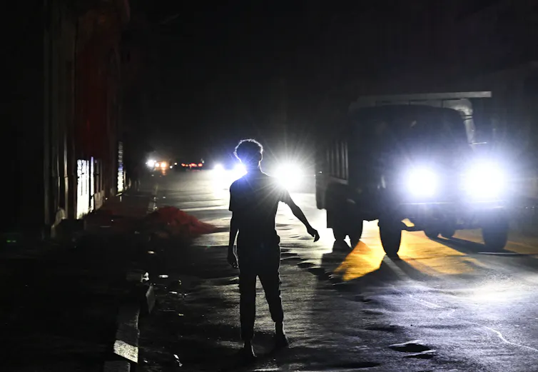 A man stands in a street with only car headlights lighting the scene.