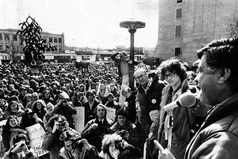 A dark-haired man talking to a huge crowd of people.