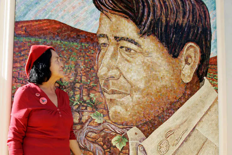 A woman with dark hair and a red dress and hat looking at a large mural of a man with brown hair.