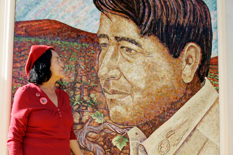 A woman with dark hair and a red dress and hat looking at a large mural of a man with brown hair.