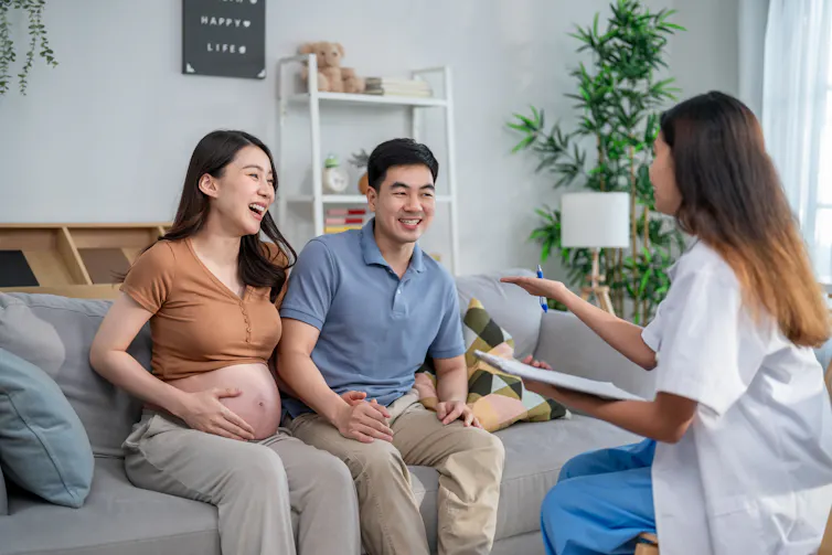 A smiling man and a pregnant woman sit on their couch while speaking to a female health visitor.