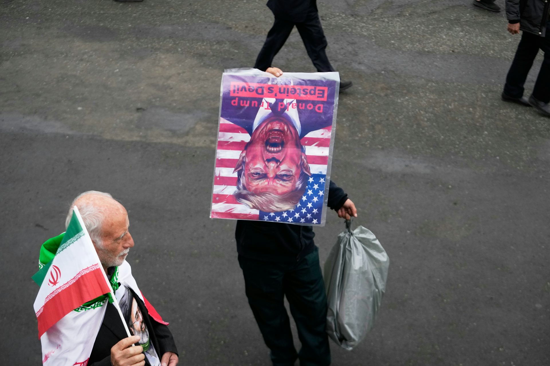 A man in the street holding an upside down photo of Donald Trump; another man beside him holds an Iranian flag.