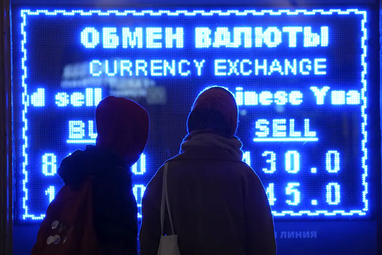Two people, seen from behind, look at a digital screen displaying an exchange rate