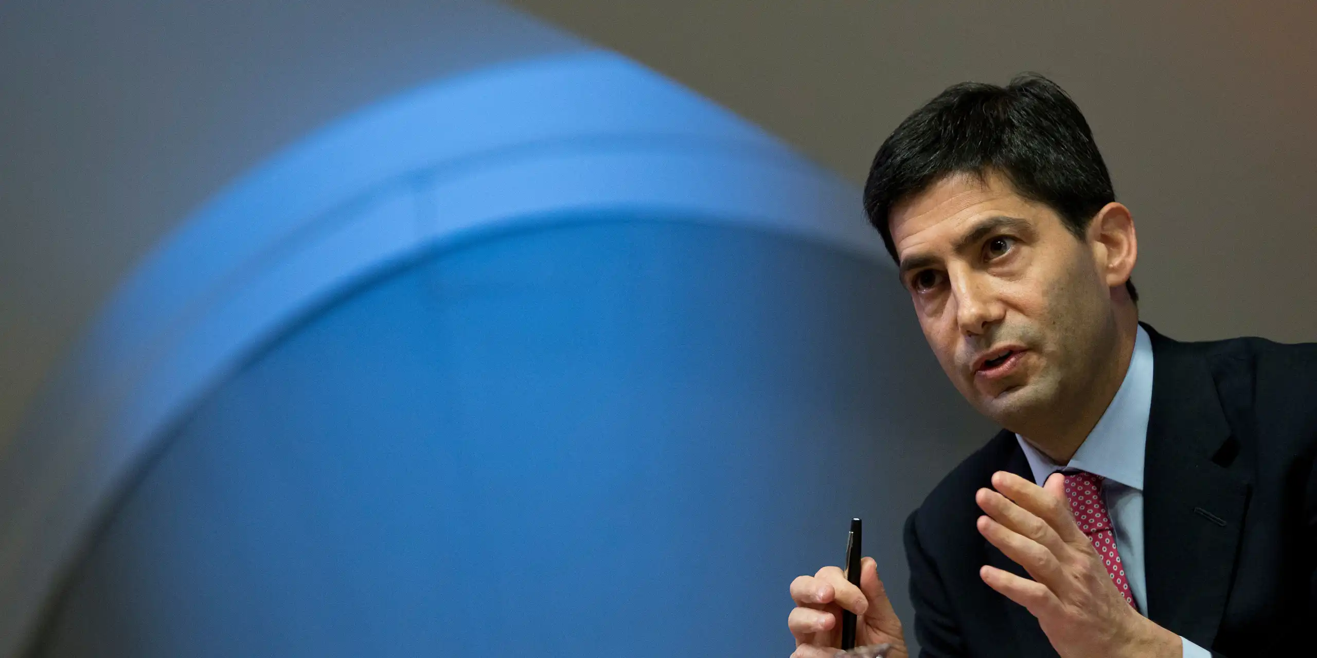 a white man with black hair in a suit gestures with his hands