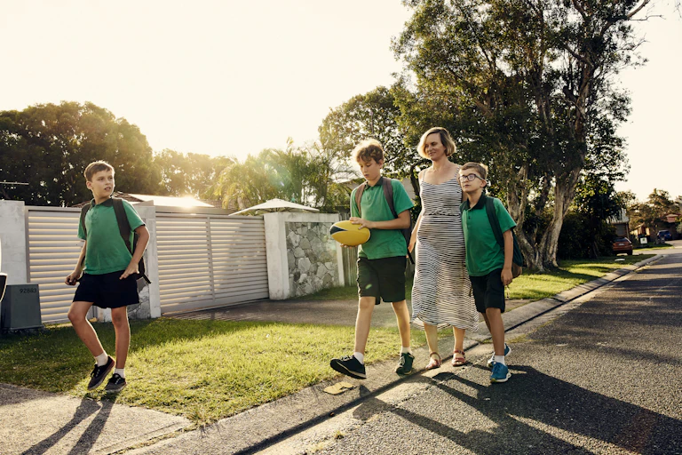 A woman walks along a suburban street with three children in school uniform.