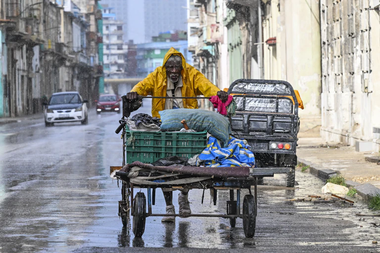 A man in a raincoat pushes a large trolley down a Cuban street in the rain