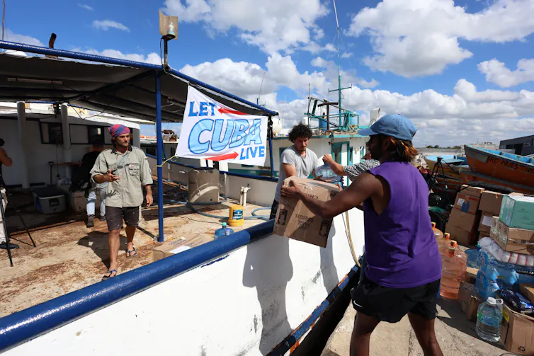 People load up a boat with supplies in front of a sign that says let Cuba live.