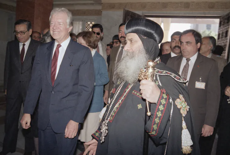 A grey-haired man in a grey suit and red tie walks beside another in a priestly robe and long flowing beard, with an entourage behind them.