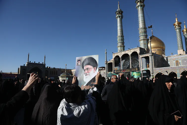 Several women in burqas stand before a shrine with tall minarets, holding a large photo of Ayatollah Khamenei.