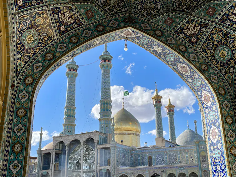 A mosque with a shining dome and tall minarets is seen through an archway with colorful intricate patterns.
