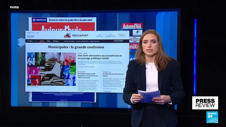 A French TV presenter in front of a screen showing headlines after March municipal elections.