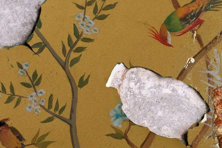 Old wallpaper showing birds in delicate branches of a tree is peeling, revealing cemenrt beneath it.