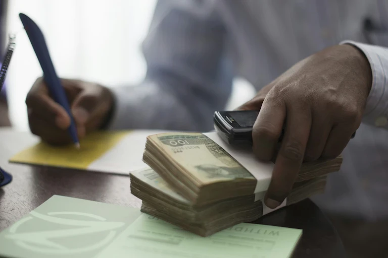 An African man's hands working at a desk, with two piles of cash in one hand he writes on a notepad with the other.