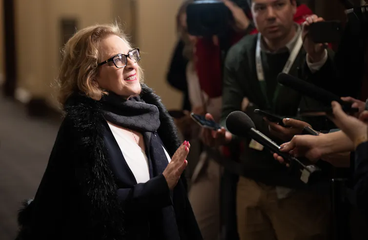 Una mujer con cabello rubio oscuro y gafas sonríe mientras habla ante los medios de comunicación.