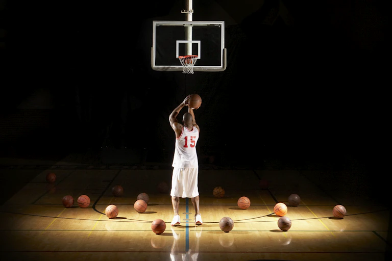Wide shot of basketball player shooting a free throw under a spotlight highlighting other basketballs surrounding him on the court