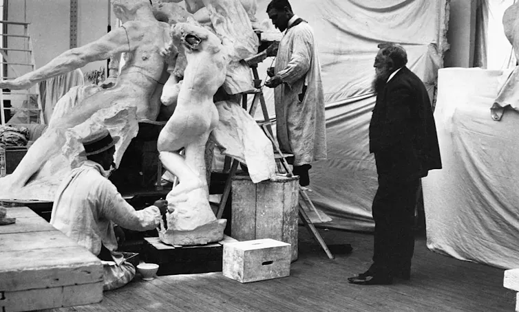 Black and white photo of bearded man wearing suit watching two men work on white sculptures.