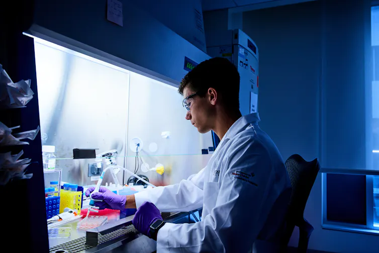 young man in white lab coat works with materials under a lab hood
