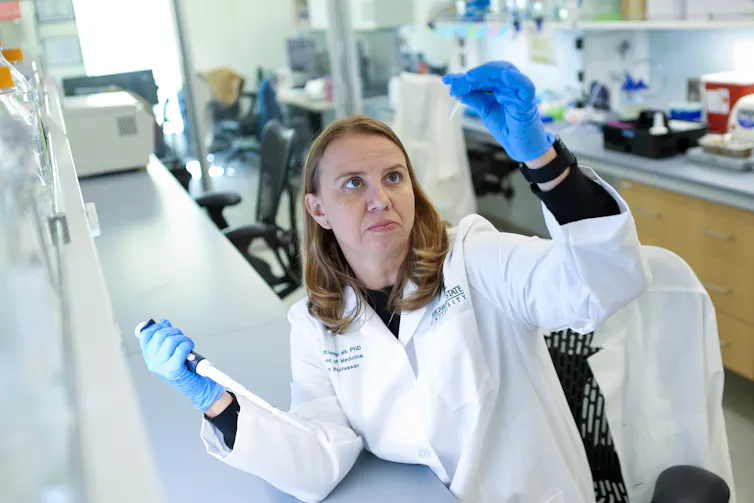 woman in white lab coat has a pipette in one hand and holds up a test tube