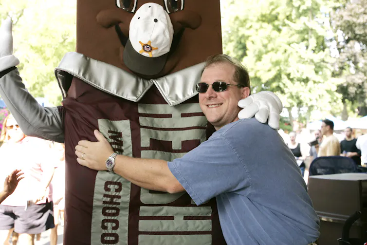 Man in blue T-shirt and sunglasses hugs a chocolate bar mascot