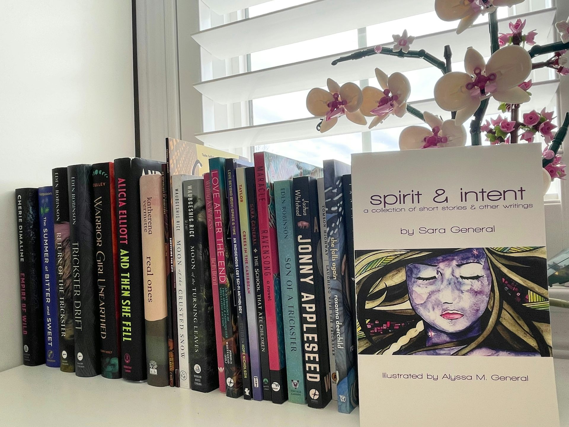 A bookshelf on a windowsill with an orchid and one book displayed with what looks like a female face and the words spirit & intent.