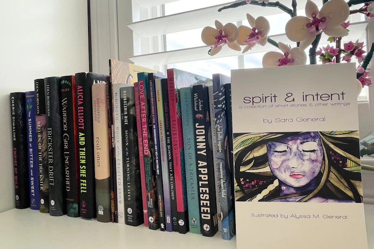 A bookshelf on a windowsill with an orchid and one book displayed with what looks like a female face and the words spirit & intent.