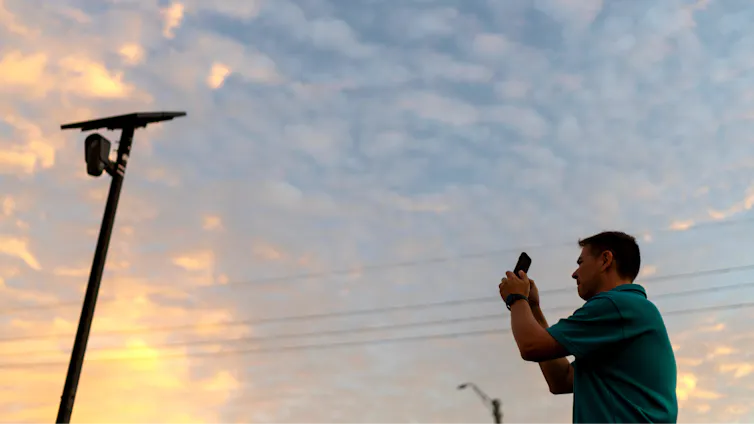 Young man wearing a polo shirt uses his smartphone to take a photograph of a camera affixed to a poll at dusk.