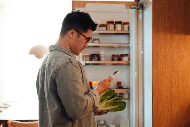Man holds bok choy in front of an open fridge door while looking at his phone