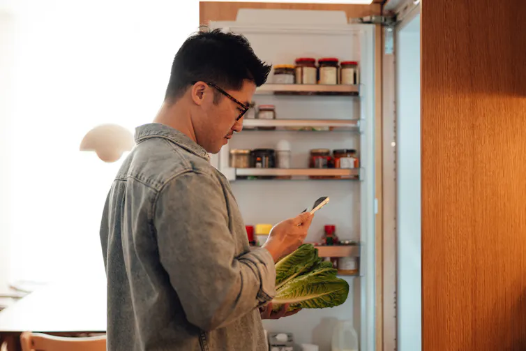 Man holds bok choy in front of an open fridge door while looking at his phone