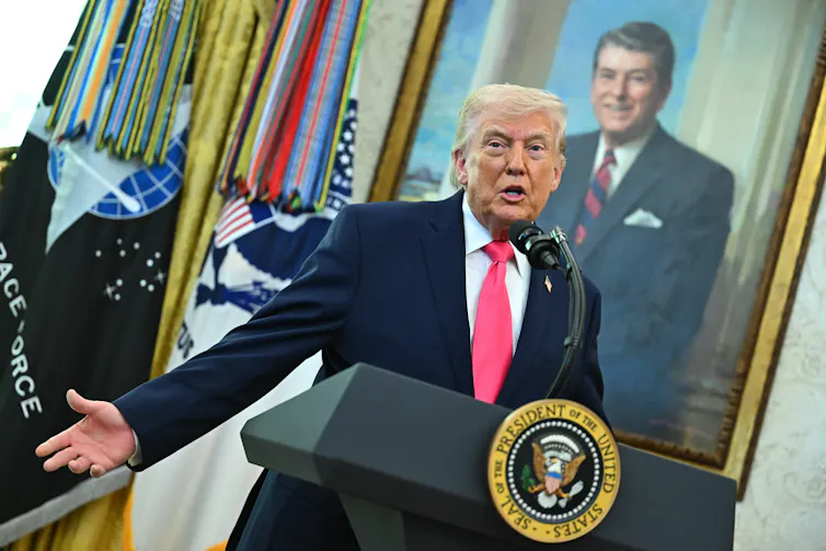 Donald Trump stands at a podium and speaks in front of a large portrait of Ronald Reagan.
