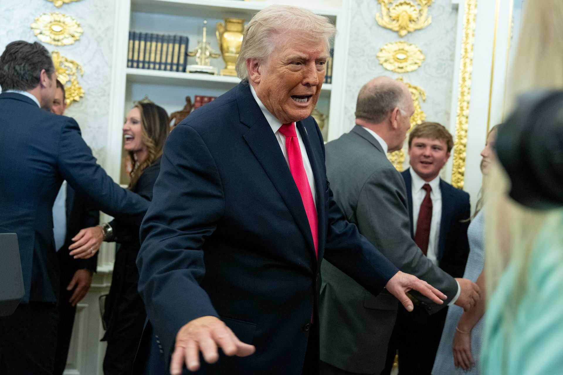 un hombre mayor con una peineta dorada y piel naranja con un traje oscuro y una expresión preocupada en su rostro. Está parado en una habitación con muebles dorados brillantes.