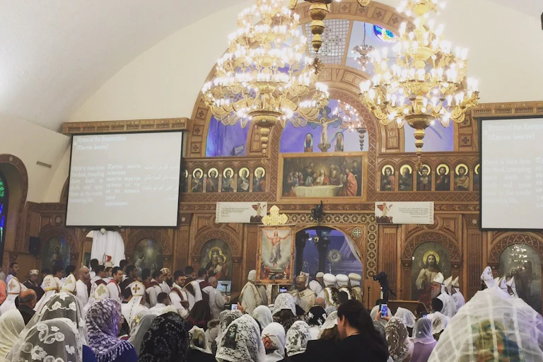 A congregation gathers for a church service, facing an altar adorned with images of Jesus, where priests in white robes stand conducting the service.