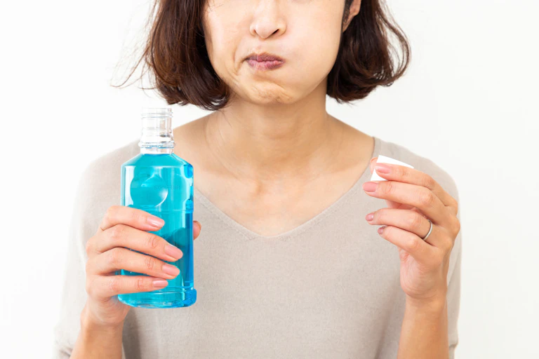 A woman holds a bottle of blue mouthwash in one hand and the cap in the other hand. She is swishes the mouthwash around in her mouth.
